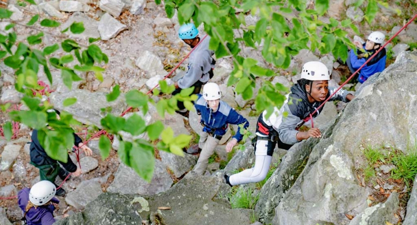 From above, a group of people wearing safety gear rock climb. 